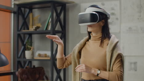 Young-woman-designer-in-office-in-virtual-reality-helmet-with-hands-makes-movements-imitating-the-work-of-graphic-interface.